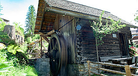 Ein traditionelles Holzhaus mit einem Wasserantrieb. Umgeben von grüner Vegetation und Bäumen.