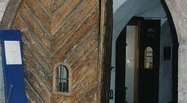 Eine große Holztür mit einem schmalen Fenster öffnet sich zu einem gewölbten Durchgang. Im Hintergrund sind weitere Türen sichtbar.