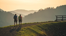 Zwei Wanderer stehen auf einem Weg und genießen die Aussicht auf die Berge. Die Atmosphäre ist friedlich, mit einem sanften Sonnenuntergang im Hintergrund.
