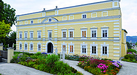 Schloss Mattighofen in Mattighofen: Ein großes, gelb gestrichenes Gebäude mit einer aufwendigen Fassade, umgeben von üppigen Blumenbeeten und grünen Rasenflächen. Das Gebäude hat mehrere Stockwerke und Balkone sowie weiße Säulen und dekorative Elemente an der Fassade. Die Umgebung wirkt gepflegt und lädt zum Verweilen ein.