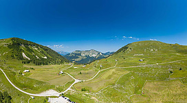 Eine weite, grüne Berglandschaft unter einem klaren blauen Himmel. Kleine Straßen schlängeln sich durch die sanften Hügel.