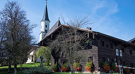 Zoll- und Heimatmuseum in Perwang am Grabensee: Ein historisches Gebäude mit charakteristischem Kirchturm, umgeben von Bäumen und Grünflächen. Die Fassade des Hauses ist aus Holz gefertigt und weist traditionelle Architekturmerkmale auf. Blühende Blumen säumen den Eingangsbereich und verleihen dem Gesamtbild einen malerischen Charakter.