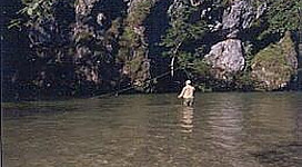 Ein Kind steht im klaren Wasser eines Baches, umgeben von hohen Felsen und Bäumen. Die Szenerie wirkt friedlich und naturnah.
