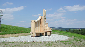 Panoramastraße in St. Aegidi: Ein großer, moderner Holzpavillon auf einer Rasenfläche, umgeben von bewaldeten Hügeln in der Ferne. Der Pavillon hat eine markante, geometrische Architektur mit Spitzen und Erker. Er scheint an einem öffentlichen Ort wie einem Park oder einer Gedenkstätte zu stehen.