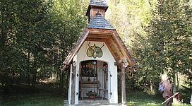 Hubertuskapelle in Seewalchen am Attersee: Das Bild zeigt ein kleines, traditionelles Gebäude mit einem spitzen Dach und Holzelementen. Es befindet sich inmitten eines waldigen Gebiets mit grüner Vegetation. Vor dem Gebäude ist eine Person zu sehen, die den Weg entlanggeht.