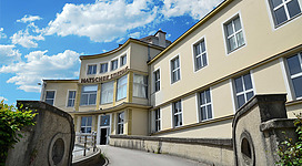 OKH - Offenes Kulturhaus Vöcklabruck in Vöcklabruck: Ein mehrstöckiges Gebäude mit einer hellen Fassade und geschwungenen Dächern. Der Eingang ist durch Treppen und Säulen gekennzeichnet. Davor erstreckt sich ein gepflasterter Vorplatz mit üppigem Grün.