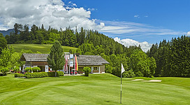 Eine gepflegte Golfanlage mit sattem grünem Rasen und einem modernen Clubhaus. Im Hintergrund sind Bäume und ein blauer Himmel mit Wolken zu sehen.