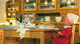 Ein gemütlicher Tisch in einer Puppenküche mit gedecktem Geschirr und einer Puppe in einem roten Kleid. Im Hintergrund sind Regale mit Geschirr und Lebensmitteln sichtbar.