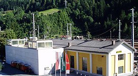 Ein modernes Gebäude mit einer gelben Fassade und mehreren Antennen steht in einer grünen Berglandschaft. Es gibt auch einige Flaggen und orangefarbene Behälter im Vordergrund.