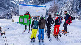Eine Gruppe von Skifahrern steht in der Schnee-Landschaft und schaut auf eine Informationstafel. Im Hintergrund sind verschneite Berge und Fichten zu sehen.