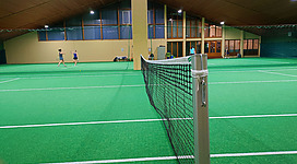 Tennishalle in Bad Kreuzen: Dieses Bild zeigt eine überdachte Tennisanlage mit einem Hartplatz in guten Zustand. Zwei Personen sind beim Tennisspielen zu sehen. Die Anlage befindet sich in einem Gebäude mit großen Fenstern und einer Holzdecke, was den Eindruck einer modernen, aber gemütlichen Sportstätte vermittelt.