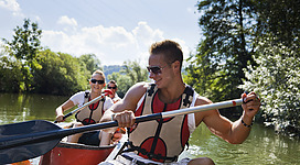 Kanu Sport Aumayr in Braunau am Inn: Eine Gruppe von Menschen in Schwimmwesten paddelt in einem kleinen Kanu auf einem Fluss durch eine grüne Landschaft mit Bäumen am Ufer. Der Mann im Vordergrund hält ein Paddel und führt das Boot an, während seine Begleiter ihm folgen.