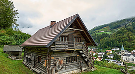 Ein traditionelles Holzhaus auf einem Hügel, umgeben von grünen Wäldern und einem kleinen Dorf im Hintergrund. Der Himmel ist teilweise bewölkt, was eine ruhige Atmosphäre schafft.