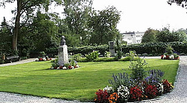 Jahnpark in Schärding: Ein gepflegter öffentlicher Park mit sorgfältig angelegten Beeten, geschwungenen Wegen und Rasenflächen. In der Mitte steht eine Statue, umgeben von üppigen Bäumen und blühenden Pflanzen. Das Bild vermittelt eine friedliche, naturnahe Atmosphäre.