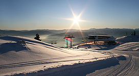Eine schneebedeckte Landschaft mit Skiliften und einem klaren blauen Himmel. Die Sonne scheint hell über den Bergen.