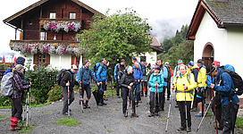 Eine Gruppe von Wanderern steht bereit am Anfang eines Wanderwegs. Im Hintergrund sind traditionelle Häuser und etwas Natur zu sehen.