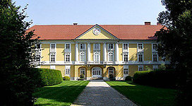 Schloss Starhemberg in Eferding: Ein gelbes zweistöckiges Gebäude mit roten Ziegeldächern und einer dekorativen Fassade. Mehrere symmetrische Fenster und ein Haupteingang mit Säulen schmücken die Vorderseite. Das Gebäude ist von üppigem Grün umgeben, mit Bäumen und einem gepflegten Rasen.