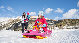 Ein fröhliches Kind fährt mit einem Schlitten auf dem Schnee. Im Hintergrund steht eine Frau mit einem kleinen Kind.