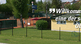 Tennisplatz des ETV Enns "1874" in Enns: Eine Tennisanlage mit mehreren Tennisplätzen befindet sich inmitten einer grünen, naturnahen Umgebung. Einige Bäume und Sträucher säumen die Anlage. Auf einem der Plätze sind Personen beim Tennisspielen zu sehen.