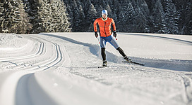 Ein Skifahrer in orangefarbener Kleidung fährt durch eine schneebedeckte Landschaft. Im Hintergrund sind verschneite Bäume zu sehen.