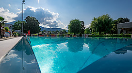 Ein klarer, blauer Swimmingpool unter einem sonnigen Himmel. Im Hintergrund sind Bäume und Wolken zu sehen.
