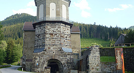 Ein historisches Gebäude mit einem runden Turm und einer steinernen Fassade. Umgeben von grünen Bergen und einer ruhigen Straße.