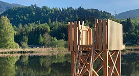 Eine Holzplattform steht an einem ruhigen See mit einer grünen Landschaft im Hintergrund. Die Plattform hat eine Treppe und einen kleinen Aussichtspunkt.