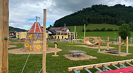 Ein Spielplatz mit verschiedenen Klettergeräten und Spielstationen. Im Hintergrund sind grüne Hügel und ein Gebäude zu sehen.