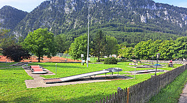 Minigolfplatz Unterach in Unterach am Attersee: Das Bild zeigt eine malerische Landschaft mit einer Parkanlage. Im Vordergrund ist ein Gelände mit Sitzbänken und Spielgeräten zu sehen, umgeben von üppigen Bäumen und Sträuchern. Im Hintergrund erheben sich steil aufragende Berge, die der Szene eine beeindruckende Kulisse verleihen.