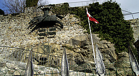 Kriegsmahnmal in Schärding: Eine Steinmauer mit einem eisernen Geländer ist zu sehen. Darauf befindet sich eine Bronzeskulptur eines Tieres. Eine Flagge ist an der Mauer angebracht. Die Umgebung ist von Bäumen und Sträuchern bewachsen.