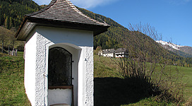 Ein kleiner, weißer Schuppen mit einem spitzen Dach steht in einer grünen Wiese. Im Hintergrund sind sanfte Hügel und Bäume sichtbar.