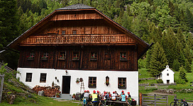 Ein traditionelles Holzhaus in einer grünen Umgebung. Eine Gruppe von Personen sitzt draußen und genießt die Natur.