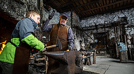 Ein Schmiedewerkstatt mit einem alten Schmied und einem Jungen. Beide arbeiten an einem Amboss, um Metall zu bearbeiten.