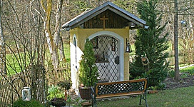Ein kleines, charmantes Gartenhaus mit einem Holzdach und Kreuz. Es steht in einem gepflegten Garten, umgeben von Bäumen und einer Holzlattenzaun.