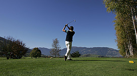 Ein Golfer schlägt einen Ball auf einem grünen Platz. Im Hintergrund sind Berge und ein klarer Himmel zu sehen.