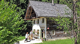 Ein traditionelles Holzhaus umgeben von Bäumen und Grünflächen. Menschen besuchen das Haus, das eine einladende Atmosphäre versprüht.