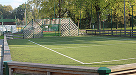 Fun Court in Andorf: Ein umzäunter grüner Tennisplatz mitten in einer waldigen Umgebung. Auf der einen Seite befindet sich ein Holzzaun mit einem abwechslungsvollen Muster aus diagonalen Linien. Dahinter sind Bäume mit dichtem sommerlichem Blattwerk zu sehen.