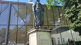 Eine Statue eines historischen Führers steht vor einem modernen Gebäude. Im Hintergrund sind grüne Bäume und eine klare Himmel zu sehen.