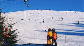 Ein malerischer Skihang mit vielen Skifahrern und einem blauen, klaren Himmel. Im Vordergrund stehen zwei Personen, die auf die Piste schauen.