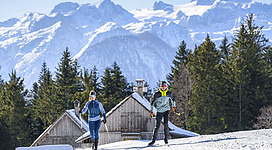 Zwei Langläufer/innen gehen nebeneinander auf einer sonnigen Loipe. Hinter ihnen ragen verschneite Holzhütten und dichte Tannenwälder auf, im Hintergrund eine imposante, weiße Bergkulisse mit Gletschern. Die Szene wirkt aktiv, klar und typisch alpin.