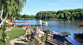 Strandbad Holzöstersee in Franking: Ein idyllischer See, umgeben von grünen Bäumen und Sträuchern. Es gibt einen hölzernen Steg mit mehreren Booten und aufblasbaren Spielgeräten im Wasser. Entlang des Ufers sind Tische und Sonnenschirme aufgestellt, die eine einladende Atmosphäre schaffen.