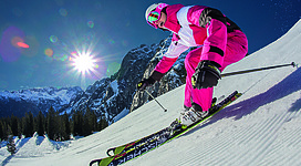 Ein Skifahrer in einem bunten Outfit fährt die verschneite Piste hinunter. Im Hintergrund sind beeindruckende Berge und ein klarer blauer Himmel zu sehen.