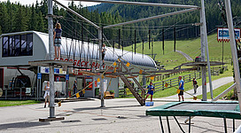 Eine Abenteueranlage mit Seilbrücken und Kletterelementen in einer alpinen Umgebung. Im Hintergrund sind Bäume und ein Gebäude zu sehen.