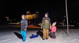 Drei Kinder stehen im Schnee bei Nacht. Sie tragen warme Kleidung und haben viel Spaß in der winterlichen Landschaft.