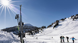 Eine schneebedeckte Landschaft mit Skiern im Vordergrund und einer Gruppe von Skifahrern im Hintergrund. Die Sonne scheint hell am klaren blauen Himmel.