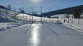 Eine glatte Eislauffläche in einer winterlichen Landschaft. Im Hintergrund sind schneebedeckte Hügel und eine klare, sonnige Skyo.