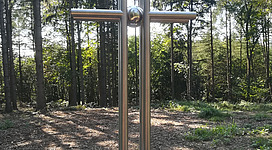 Gipfelkreuz Kürnbergwald in Wilhering: Ein metallisches Kreuz auf einem Sockel aus Stein steht in einem bewaldeten Gebiet. Das Kreuz ist schlicht und elegant gestaltet, mit einem kugelförmigen Element in der Mitte. Die Umgebung ist von dichten Bäumen und Sträuchern umgeben, die eine ruhige und natürliche Atmosphäre schaffen.