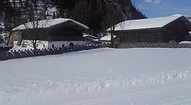 Ein winterlicher Landschaft mit schneebedeckten Feldern und traditionellen Holzhäusern. Im Hintergrund sind Berge und ein klarer blauer Himmel zu sehen.