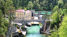 Eine historische Wasserkraftanlage umgeben von grünen Bäumen. Der Fluss fließt in lebhaftem Türkis durch die Landschaft.