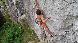 Ein Kletterer erklimmt eine steile Felswand in einer natürlichen Umgebung. Unter ihm ist grünes Gras und einige Sträucher zu sehen.